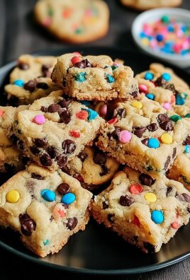 Chewy cookie bars filled with colorful M&M candies and chocolate chips stacked on a black plate
