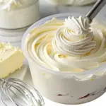 A close-up of a tub of fluffy whipped pudding frosting being swirled with a piping nozzle, surrounded by whipped cream, a wire whisk, and a block of butter.
